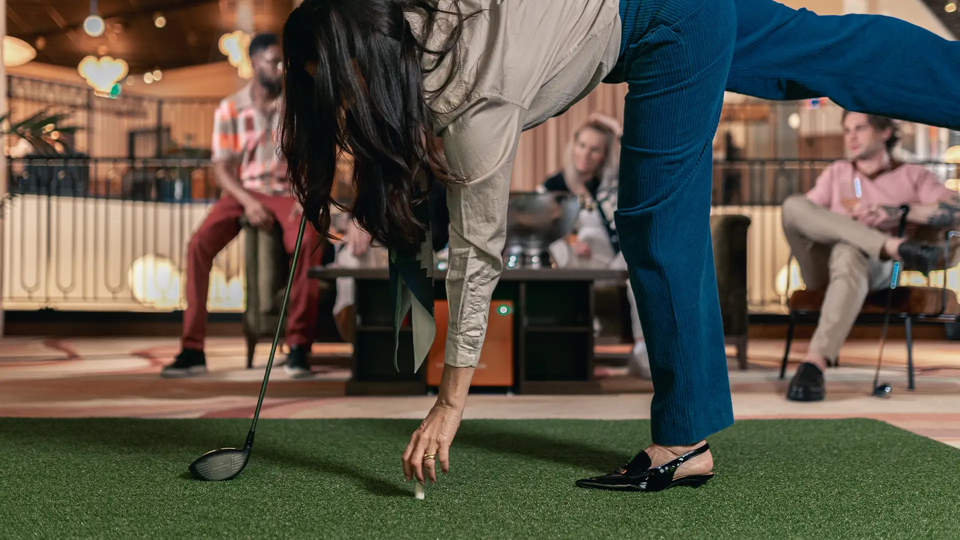 fem-kamp-goteborg Kvinna förbereder ett slag i inomhusgolf medan vänner ser på inne i en elegant lounge-miljö.